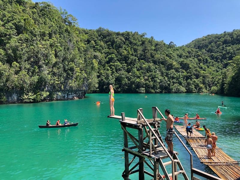 People diving and swimming at Sugba Lagoon, with kayaks, a canoe, and bamboo rafts on turquoise water surrounded by jungle covered limestone cliffs
