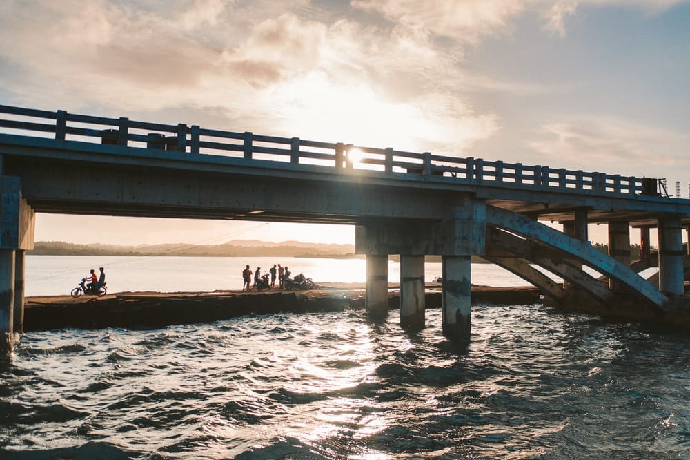 Sunset view of Catangnan Bridge with people gathered on the rocky shore below and a motorbike parked nearby, General Luna