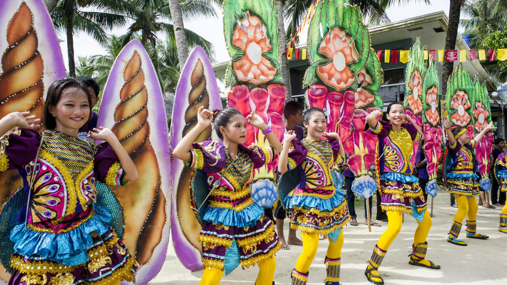 Dancers wearing colorful purple, gold, and blue costumes with giant shell and flower shaped props performing at the General Luna town fiesta in Siargao