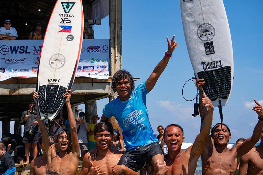 A winning surfer raised on teammates shoulders holding a peace sign, surrounded by cheering competitors and surfboards at a surf competition, Cloud 9