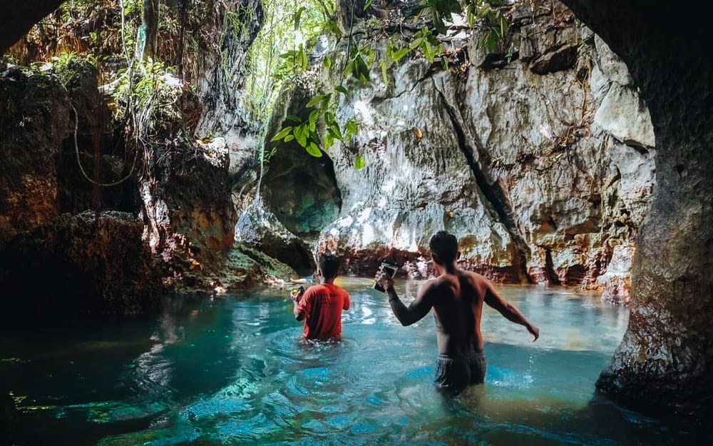 2 people wading through clear turquoise water inside Tayangban Cave Pool, surrounded by limestone rock walls and hanging jungle vines