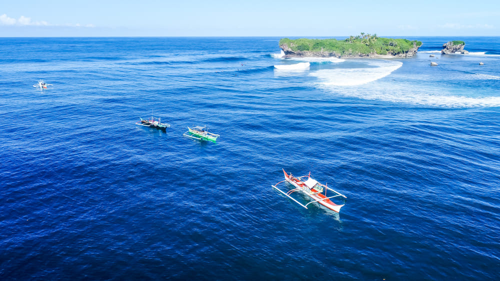 Aerial view of 3 outrigger bangka boats floating on deep blue water near a small rocky islet with waves breaking around it off Siargao