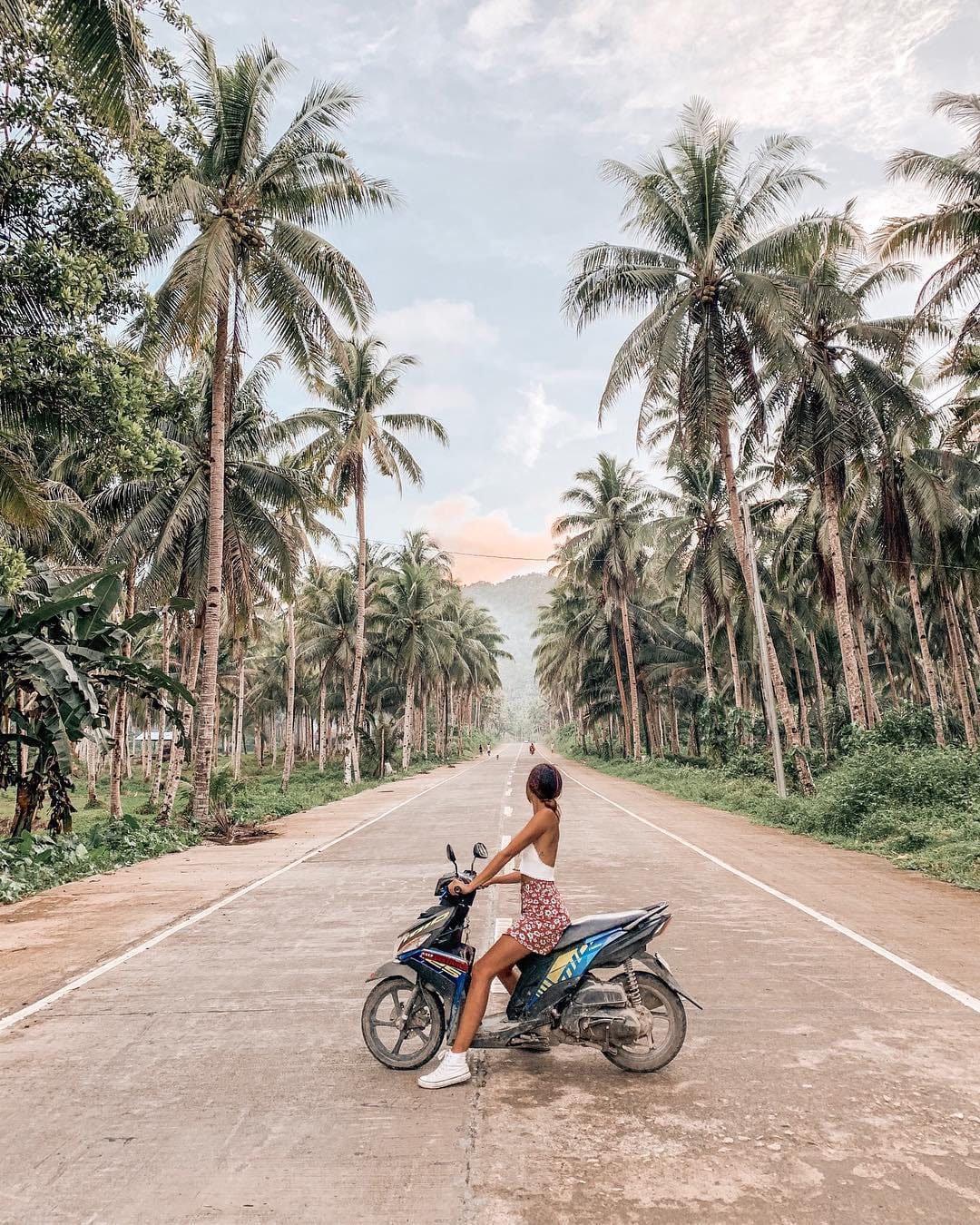 A woman sits on a parked motorbike on an empty road lined with tall coconut palm trees stretching toward distant hills on Siargao Island