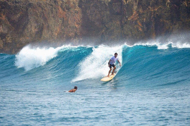 A surfer in a blue rash guard rides inside a breaking wave beside dark rocky cliffs, as another swimmer watches from the water nearby