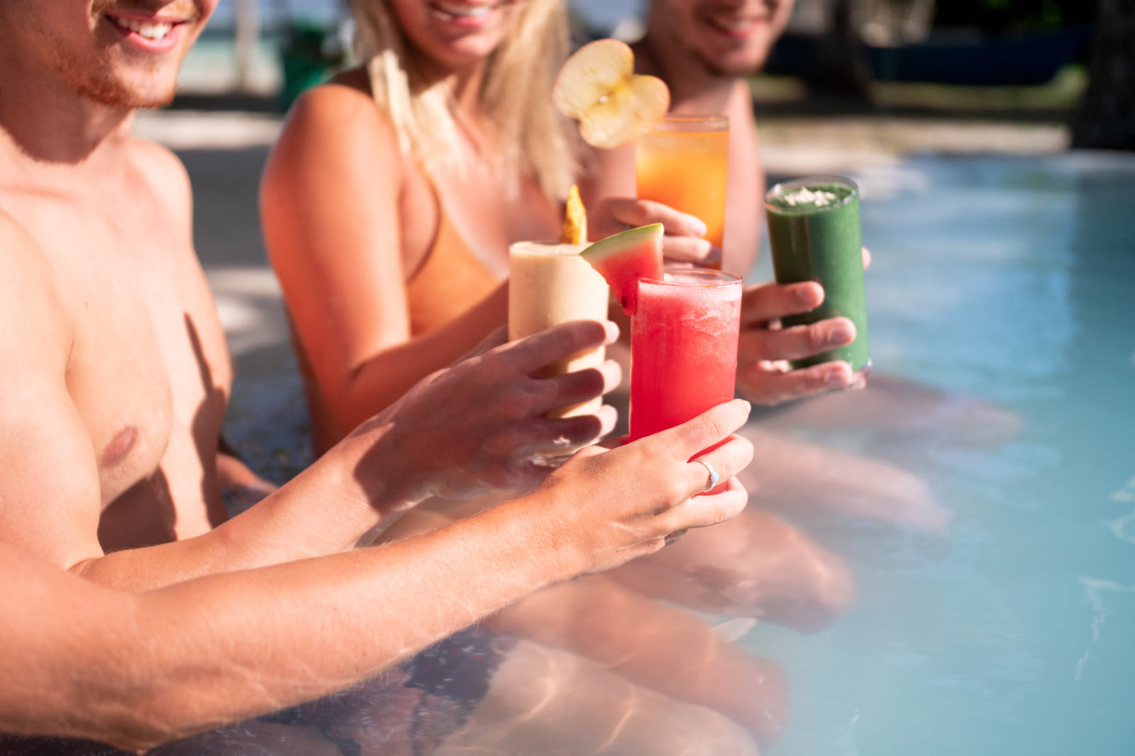 3 guests standing chest-deep in a pool at Bravo Beach Resort Siargao raise glasses of colorful fresh fruit drinks together in a toast