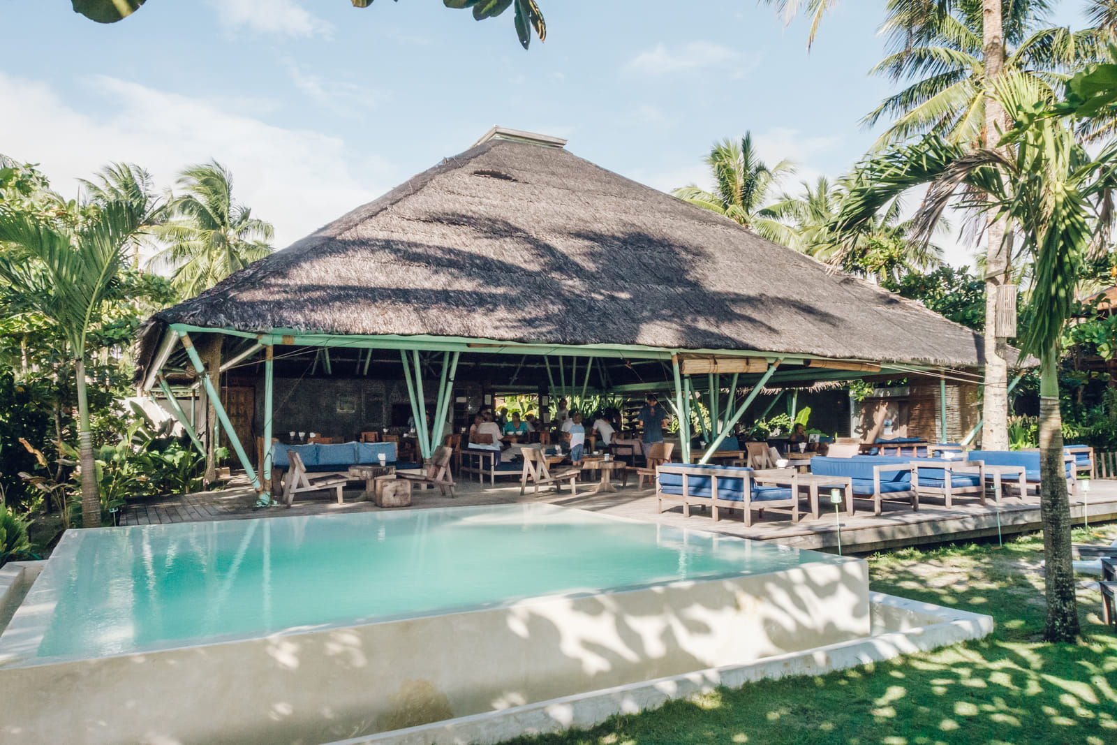 The main thatched-roof pavilion and pool at Bravo Beach Resort Siargao, with guests seated at tables and loungers, framed by coconut palms