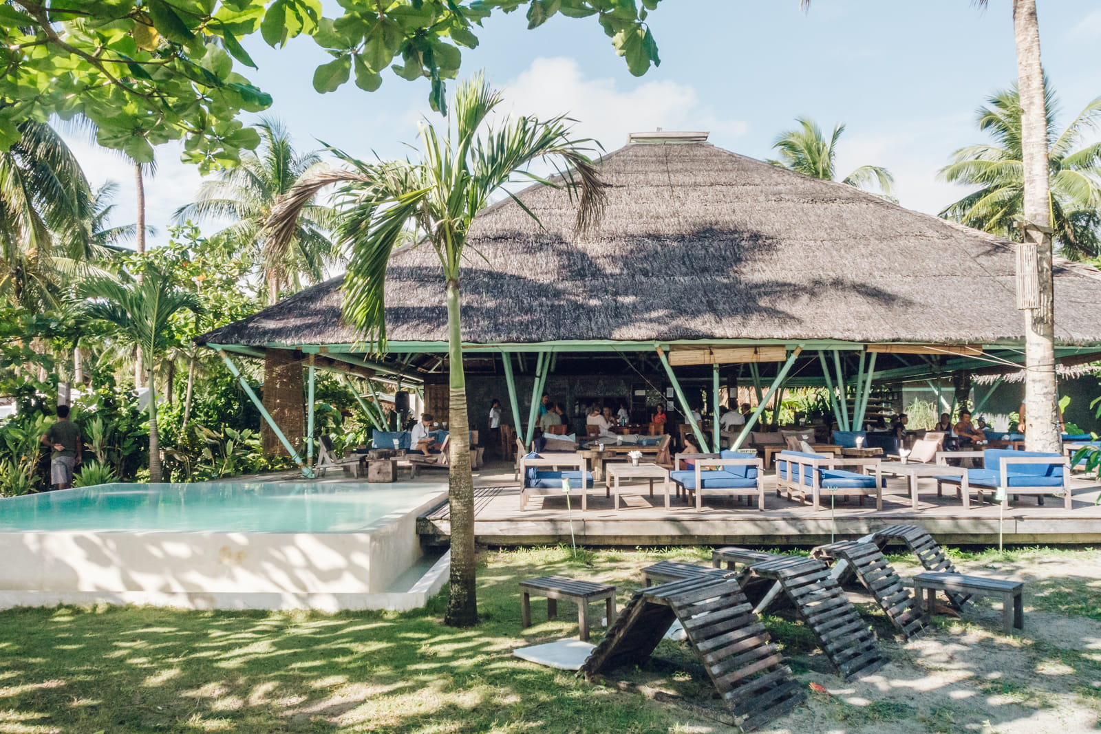 Wooden A-frame lounge chairs stacked on the lawn beside the pool and thatched pavilion at Bravo Beach Resort Siargao, in dappled shade