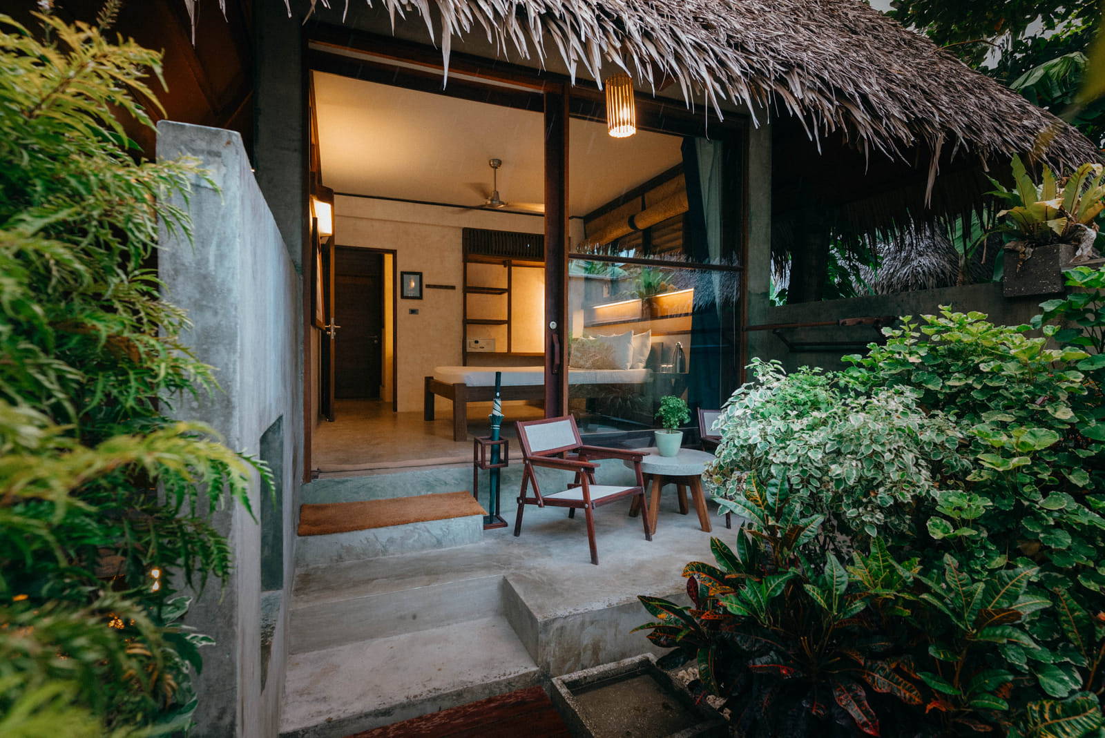The entrance to a Garden Room at Bravo Beach Resort Siargao at dusk, with a lit interior, thatched roof, and a small patio with 2 chairs