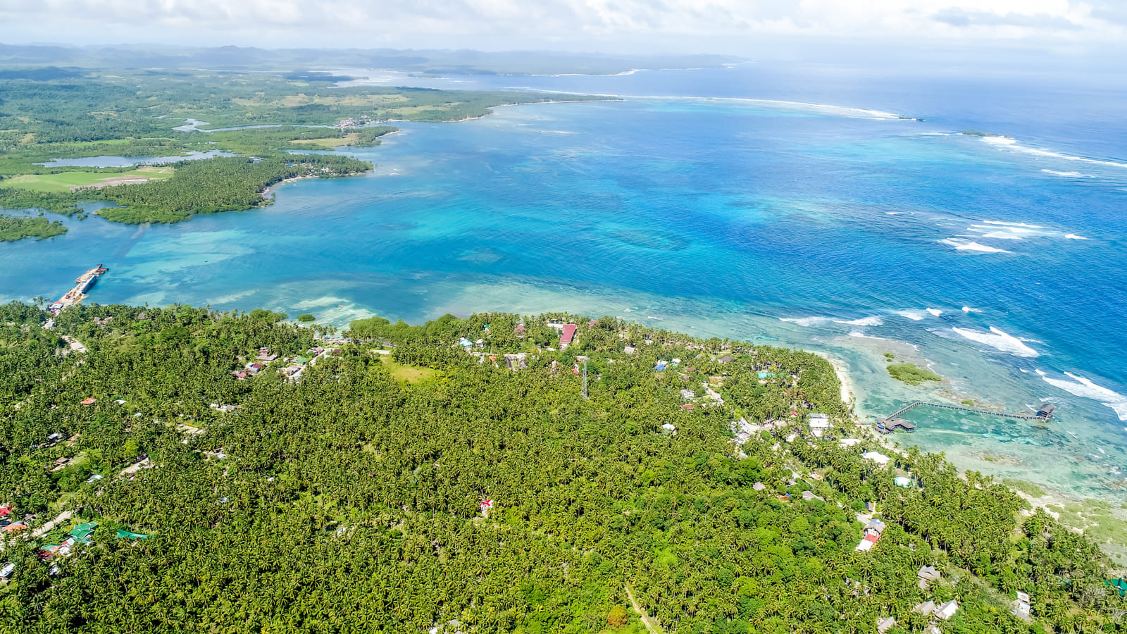 Aerial view of the palm-covered Siargao coastline with a turquoise lagoon, coral reef patches, and the open blue ocean stretching to the horizon
