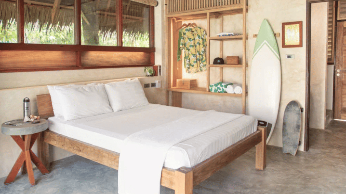 A guest room at Bravo Beach Resort Siargao with a wooden bed, white linens, a surfboard leaning against the wall, and open wardrobe shelving