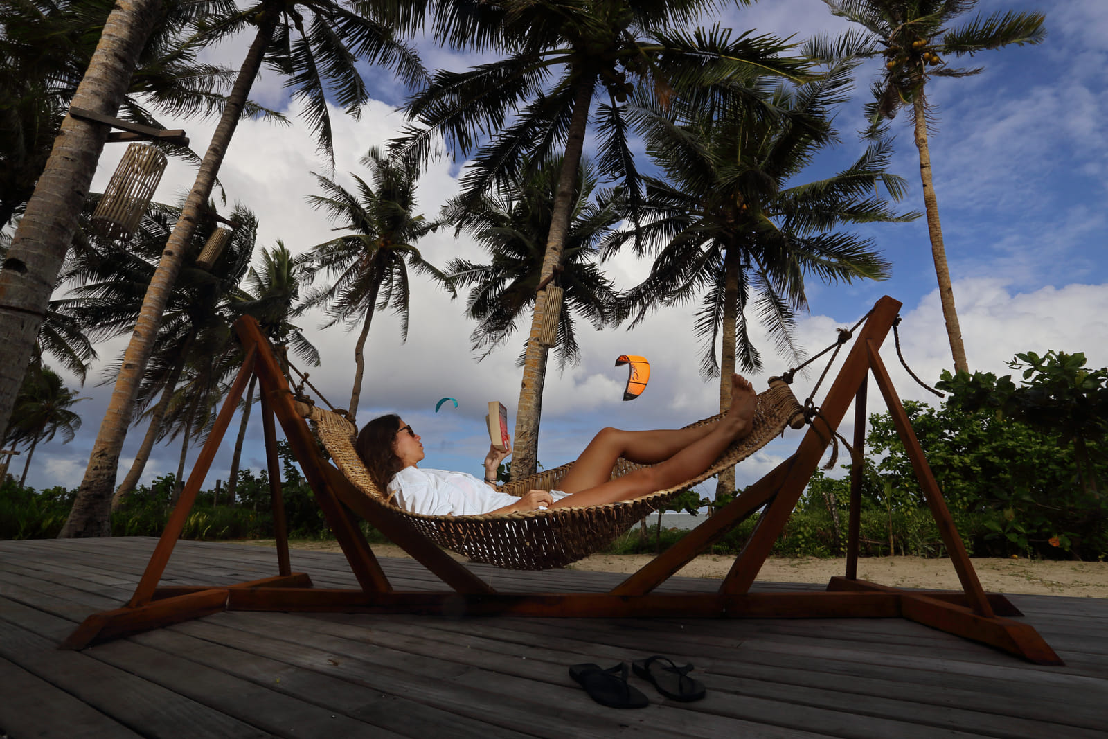 A guest relaxes reading a book in a wooden-framed hammock on a deck at Bravo Beach Resort Siargao, with palm trees and a kitesurfer in the distance