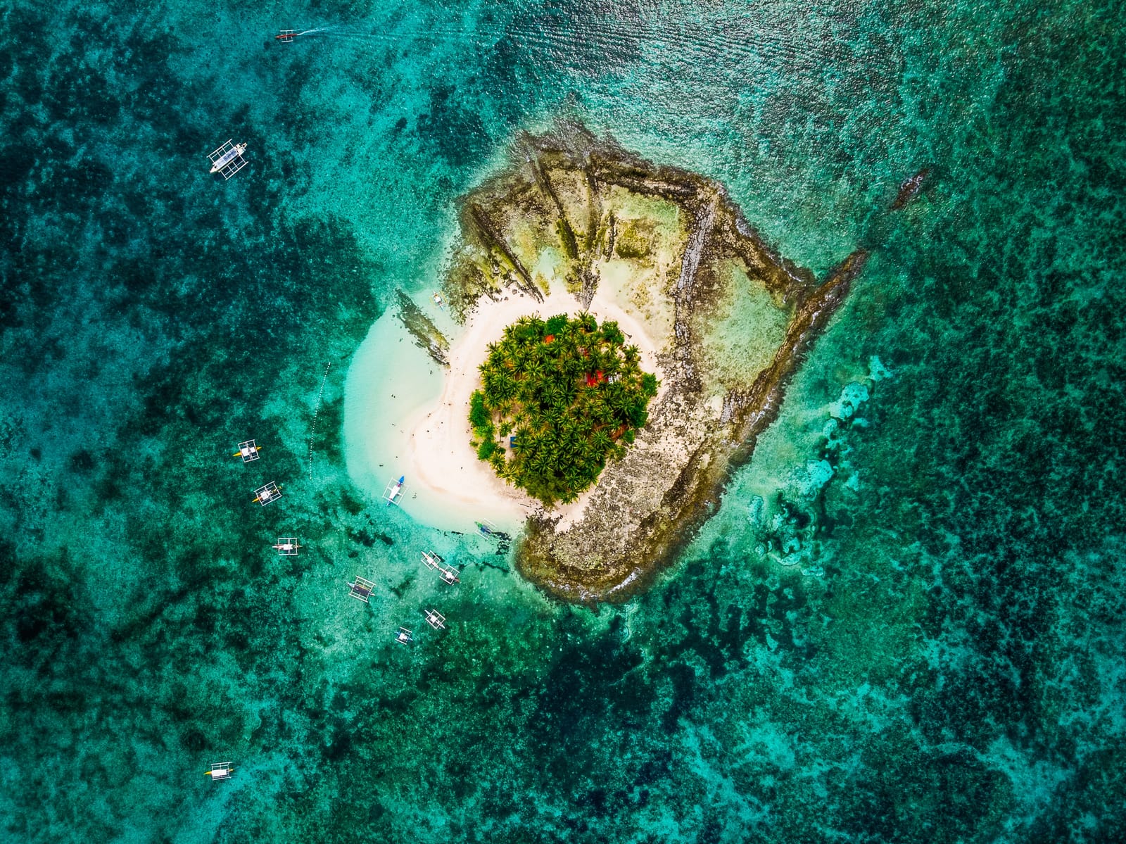 An aerial drone view of a small islet with white sand and dense green trees, surrounded by turquoise reef water and several outrigger boats