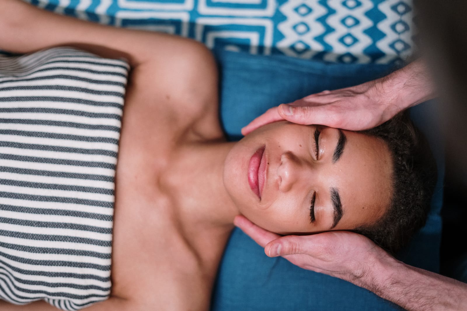 A person with eyes closed receives a facial massage, hands cradling their head while lying on a blue cushion with a geometric-patterned pillow