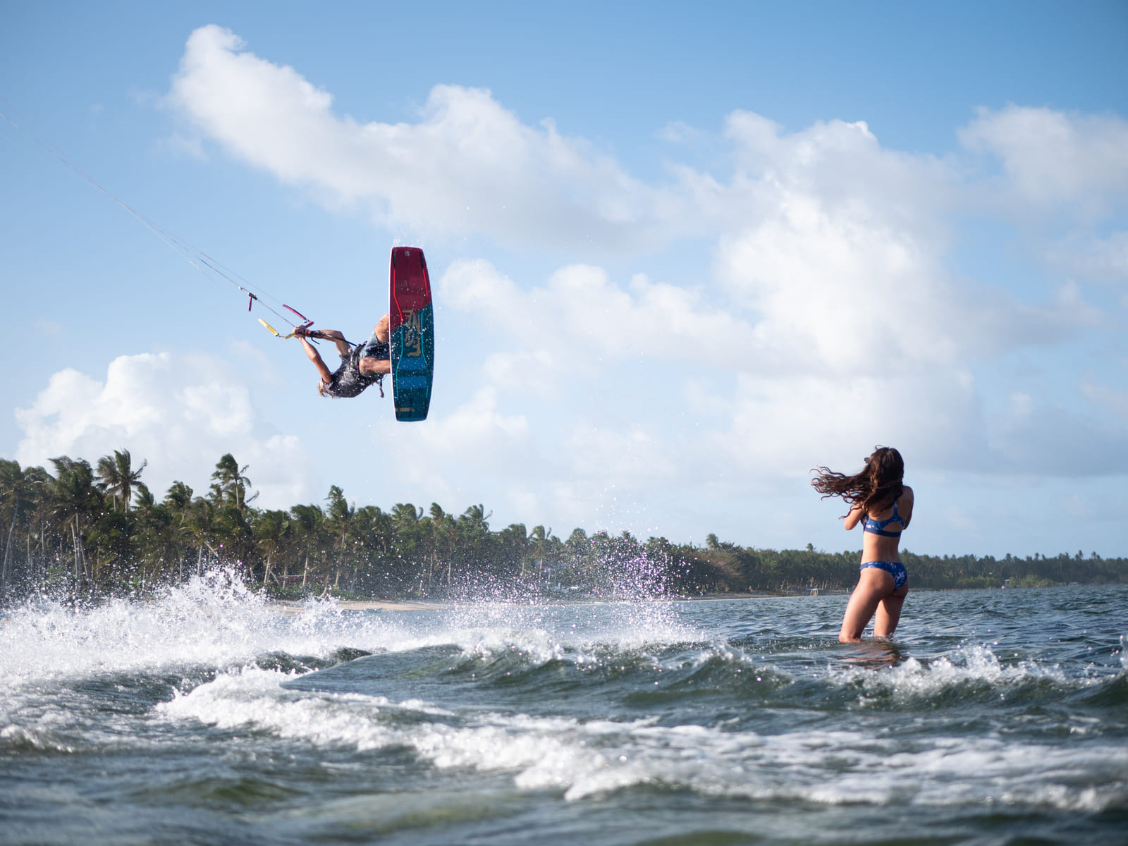 A kiteboarder launches high into the air off a wave while a woman watches from the water, with a palm-lined coastline behind them