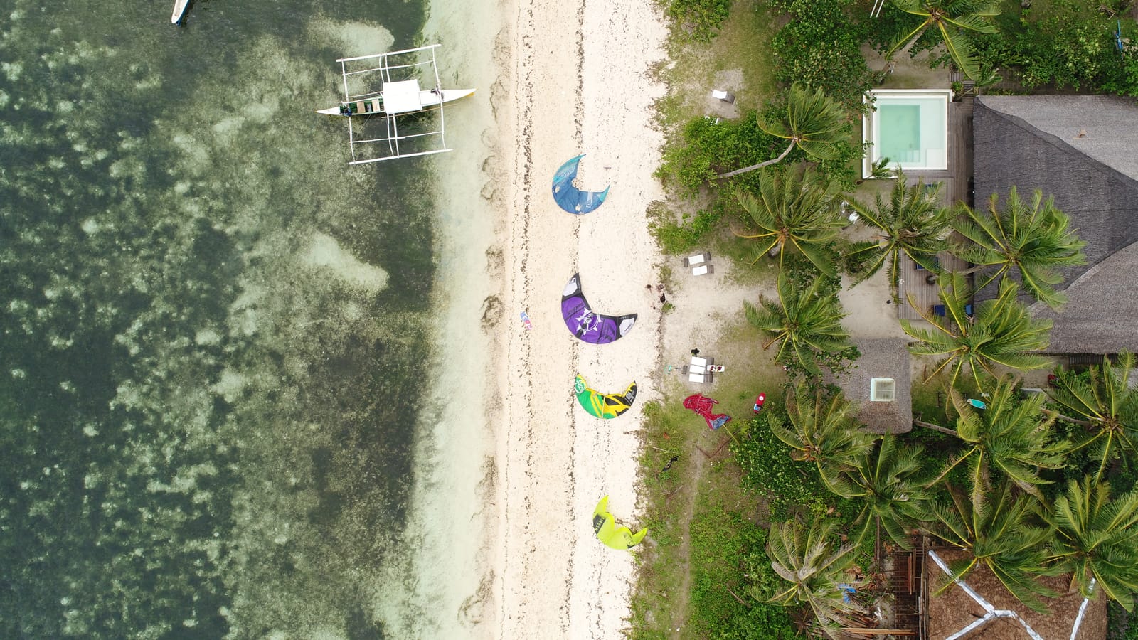 Aerial view of colorful kiteboarding kites laid out on a white sand beach in Siargao, with an outrigger boat and palm trees nearby