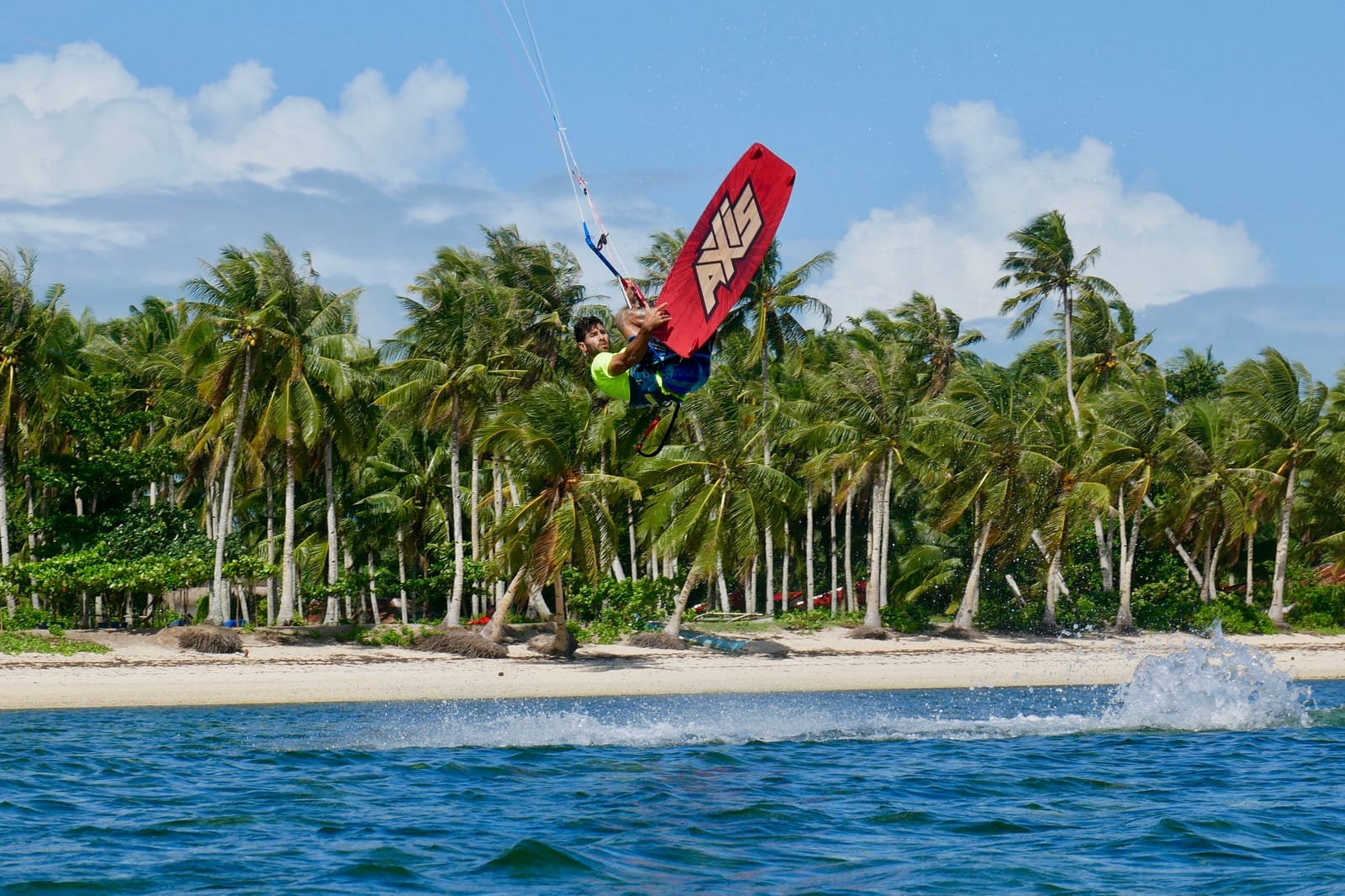 A kiteboarder jumps high above the water on a red board, with a dense palm-lined white sand beach in Siargao stretching behind him