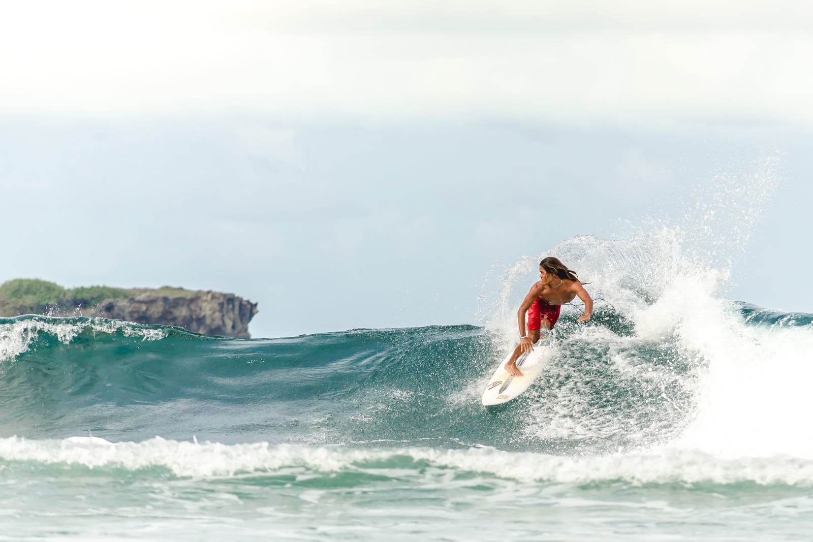 A surfer in red shorts carving a turquoise wave with white spray, a rocky islet visible on the horizon near General Luna, Siargao