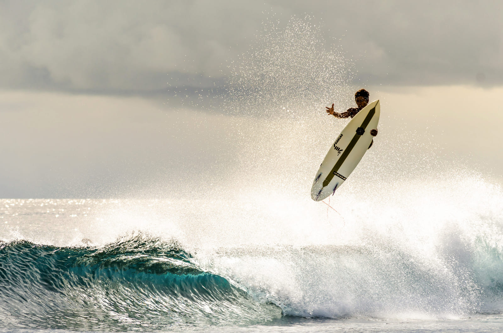 A surfer launching airborne above a breaking wave, gripping the surfboard, with sunlit spray and a dramatic cloudy sky over the ocean in Siargao