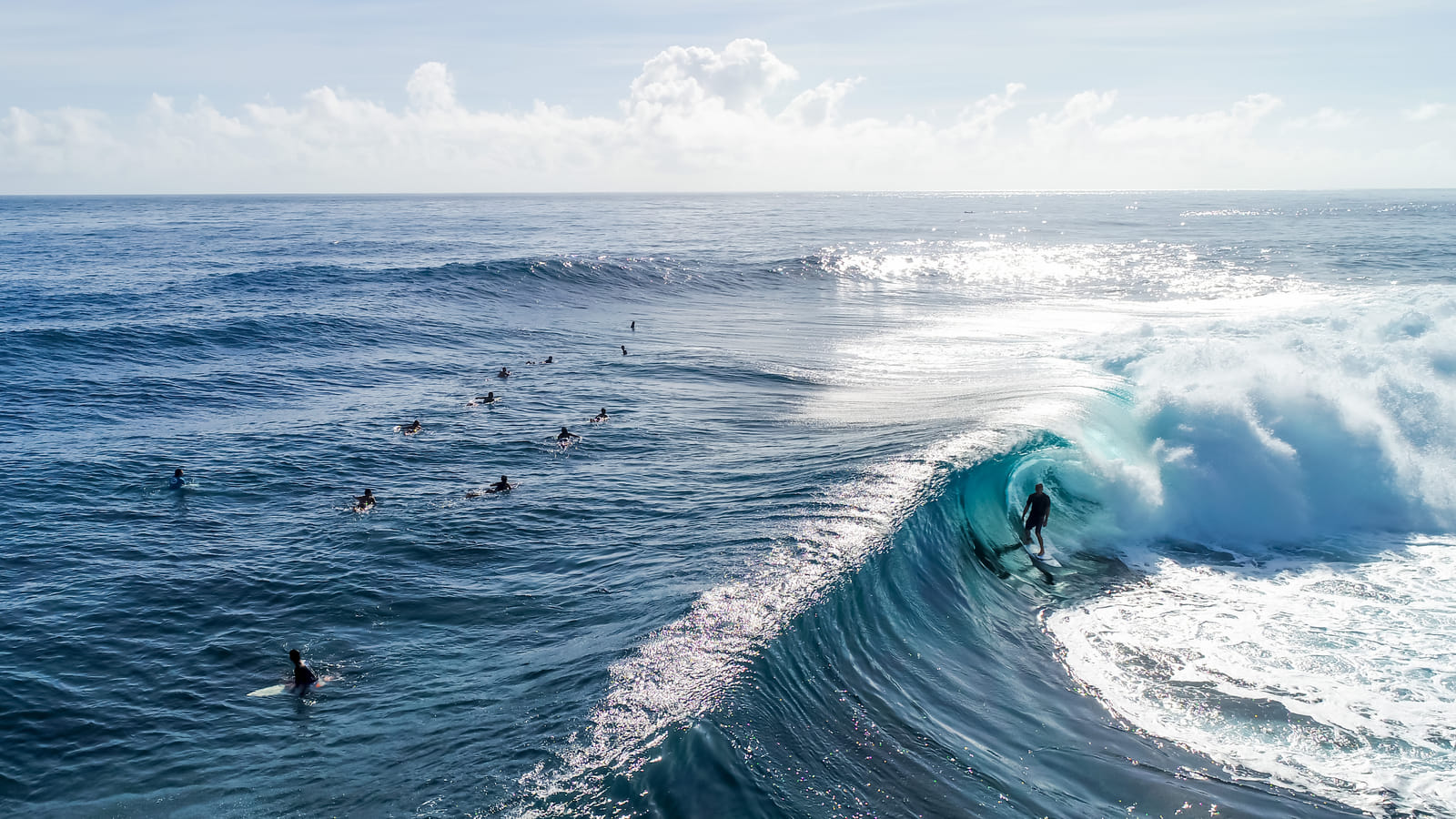 Aerial view of a lineup of surfers paddling out across glassy blue water while one rides inside a barreling wave at Cloud 9, Siargao