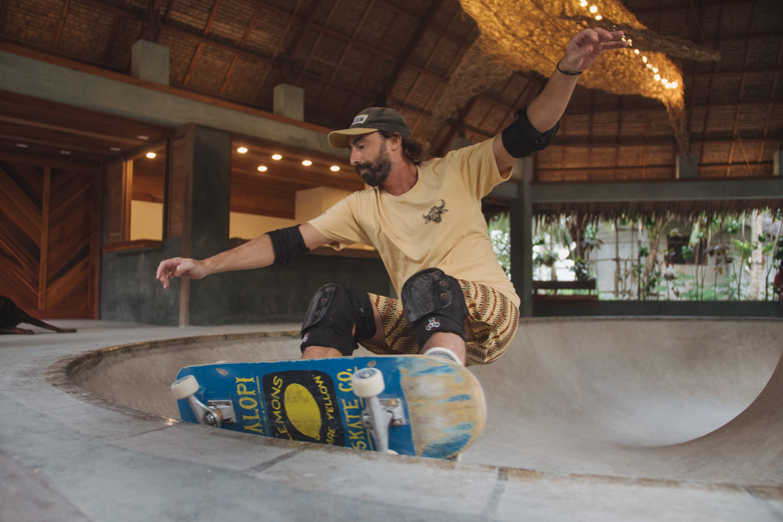 A skater in knee pads dropping into a concrete skate bowl under a thatched roof at the Bravo Skate Bowl, Bravo Beach Resort Siargao