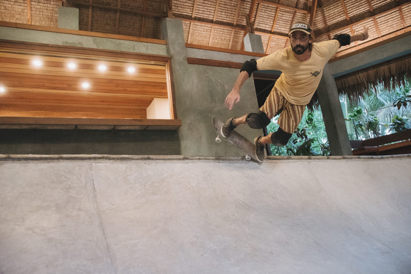 A skater with knee pads dropping into the concrete bowl beneath a thatched roof at the Bravo Skate Bowl, Bravo Beach Resort Siargao