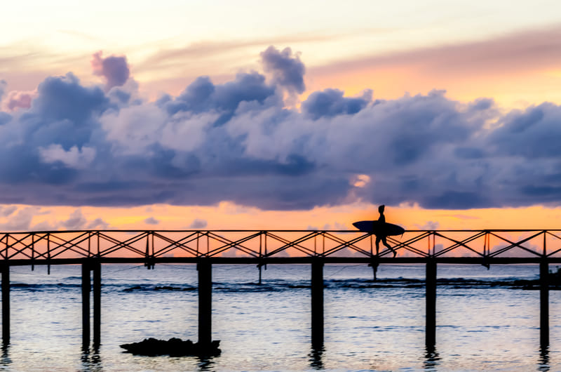 A silhouetted surfer carrying a surfboard walks across a long wooden bridge over water at sunset, with dramatic pink and purple clouds overhead