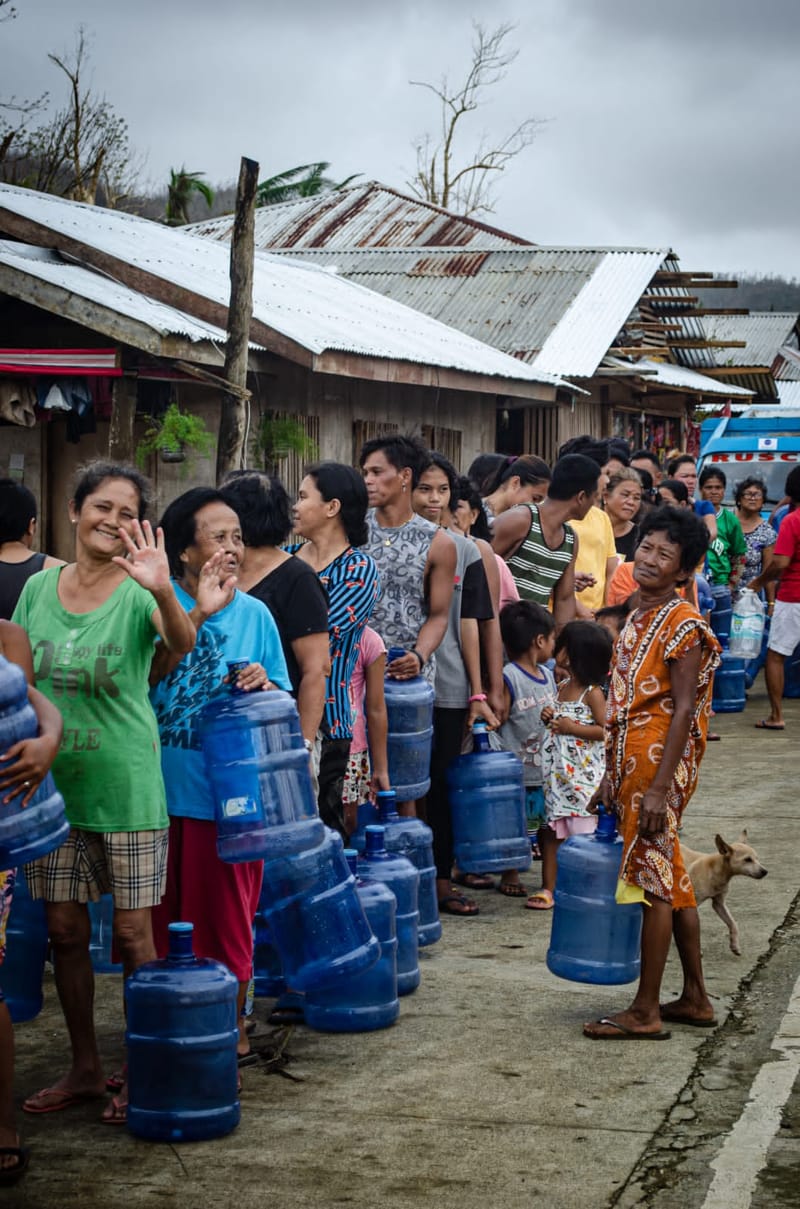 A line of residents wait with large blue water jugs beside typhoon-damaged homes with rusted roofs during a relief operation in Siargao