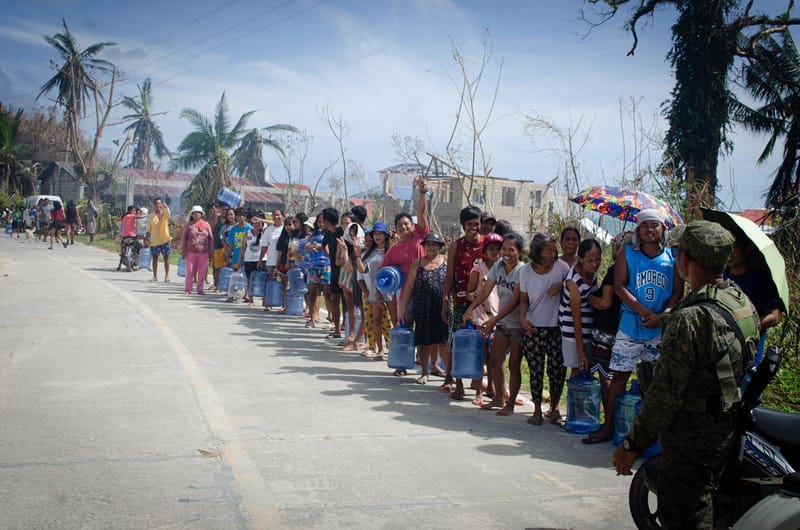 A long line of residents with water containers waits along a road lined with storm-damaged trees during a relief operation in Siargao