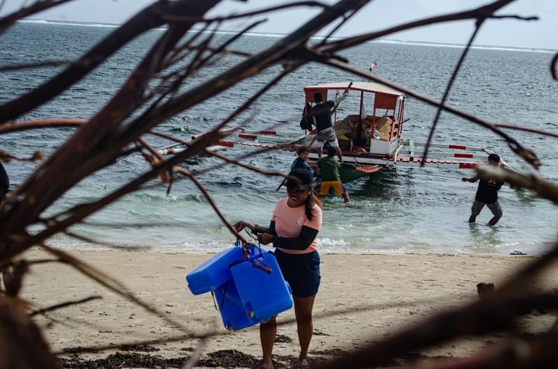 A woman carries a blue container along the shore as a boat unloads relief supplies in the background during a Siargao relief operation