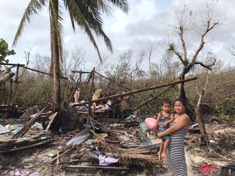 A woman holds a child beside the flattened remains of a home amid snapped trees, showing Siargao in the aftermath of Super Typhoon Odette