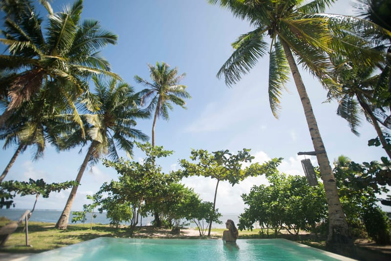 A person relaxes in an outdoor swimming pool framed by tall coconut palms and green shrubs, with the sea visible beyond, at Bravo Beach Resort Siargao