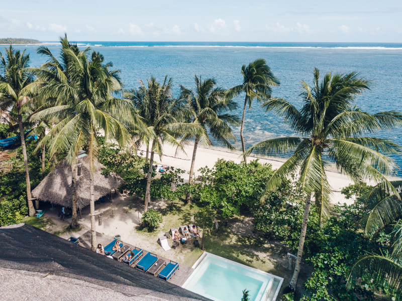 Aerial view of Bravo Beach Resort Siargao with a pool, thatched cabana, and sun loungers surrounded by palm trees beside a white sand beach and sea