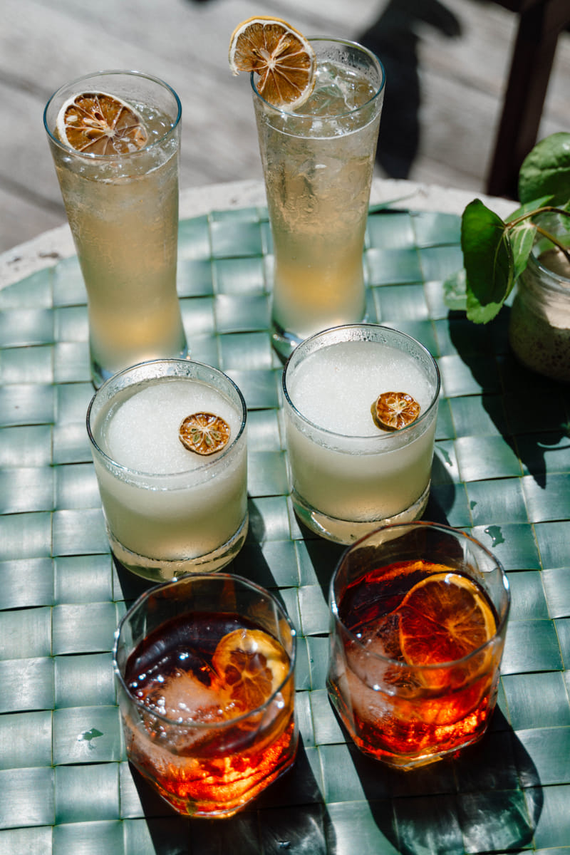Six cocktails including tall highballs with dried citrus garnish, cloudy sours, and negronis arranged on a woven-top table