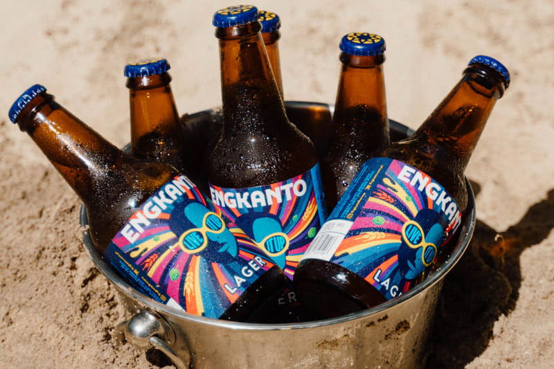 Metal bucket filled with ice and 6 chilled Engkanto Lager beer bottles with blue bottle caps, set on the sand in bright sunlight