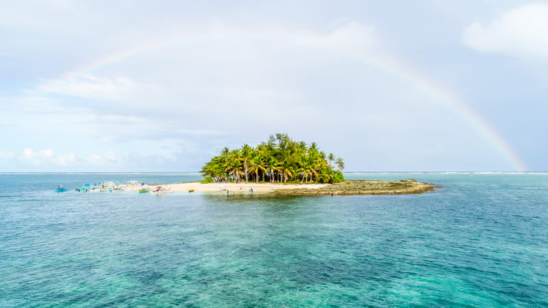 Guyam Island, a small palm-covered islet surrounded by turquoise water with outrigger boats and a rainbow overhead