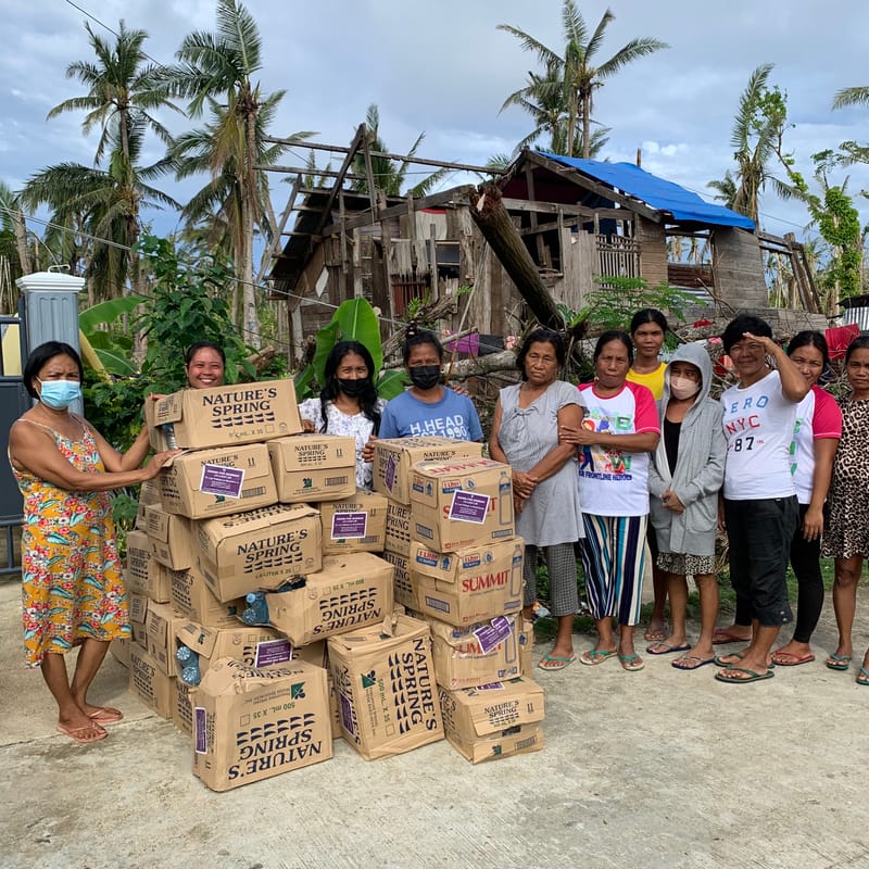 Local residents standing beside stacked relief supply boxes in front of a storm-damaged house, part of a Bravo Beach Resort Siargao relief drive