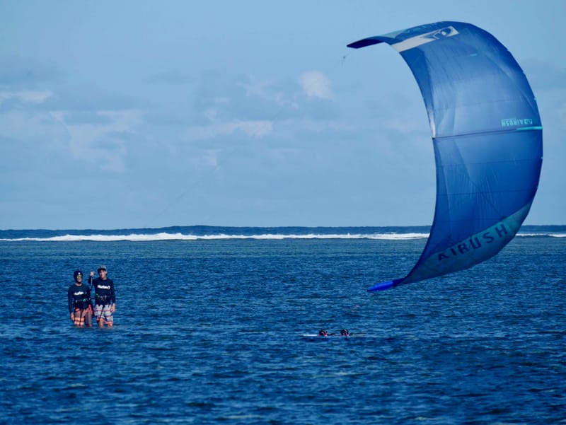 Two people wading in shallow water flying an Airush kiteboarding kite, a reef break visible in the distance