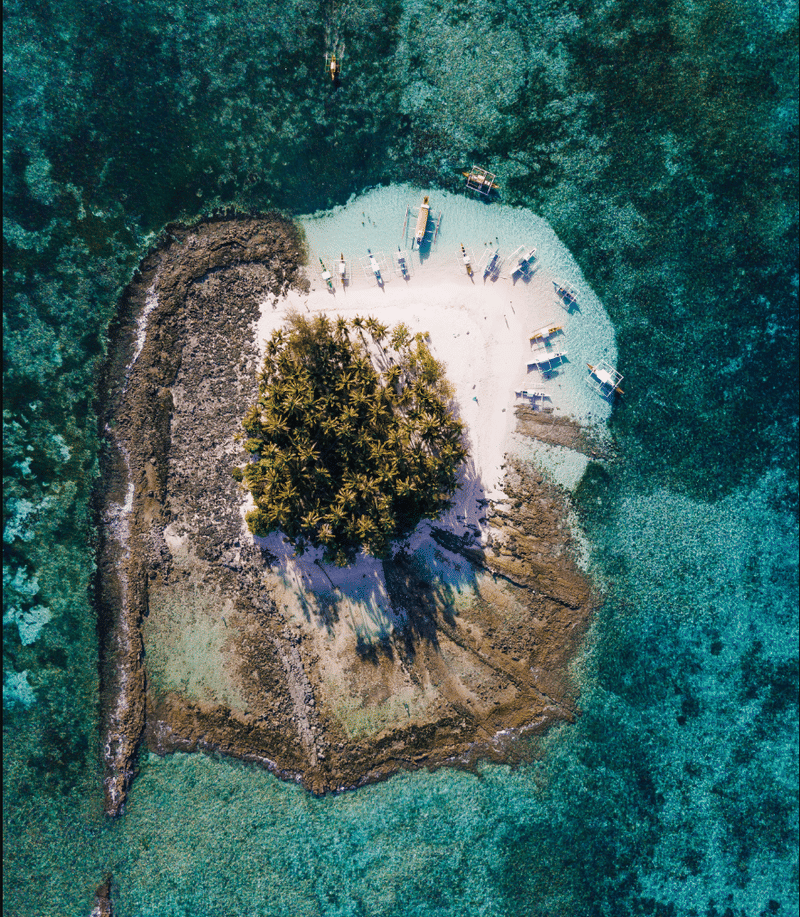 Aerial view of a small palm covered island fringed by white sand and turquoise reef, with outrigger boats moored nearby in the Philippines