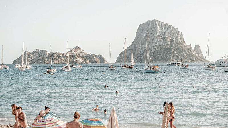 Sailboats and yachts anchored in a scenic bay with sunbathers and swimmers on the beach and a rocky island cliff on the horizon