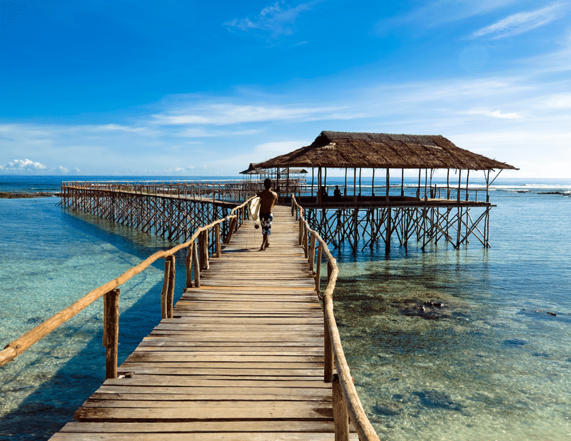 A surfer carrying a white surfboard walks along a long wooden pier leading to a thatched-roof pavilion over clear turquoise water