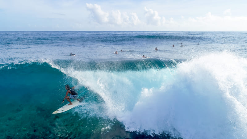 Aerial view of a surfer riding down the face of a large breaking wave, with several other surfers waiting in the lineup off Siargao
