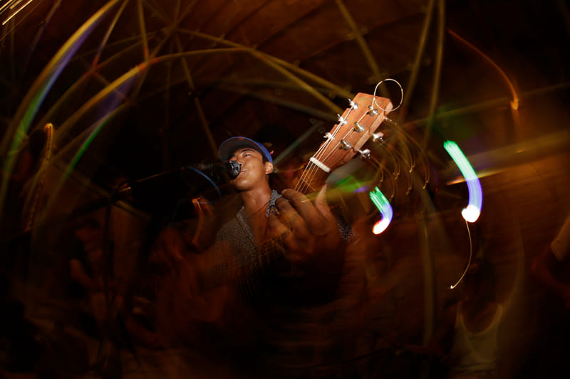 A musician sings into a microphone while holding an acoustic guitar, captured with dramatic light trail effects at a night performance
