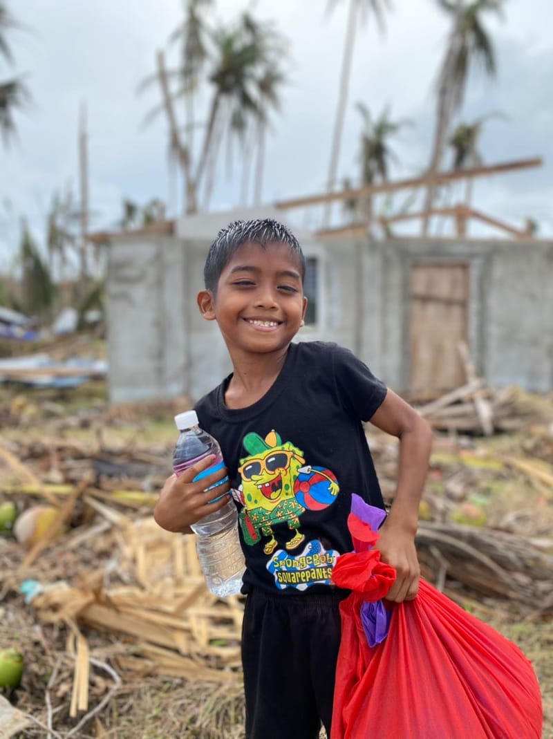 A smiling boy holds a bottle of water and a red relief supply bag in front of a storm-damaged building during a Siargao relief operation