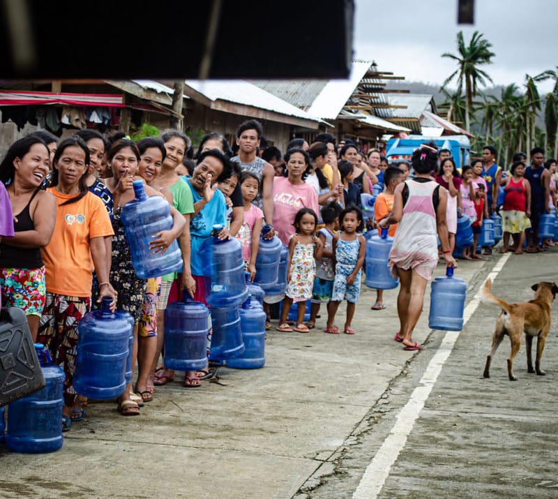 Women and children line up with blue water containers along a street of storm-damaged homes with rusted roofs during a relief operation in Siargao
