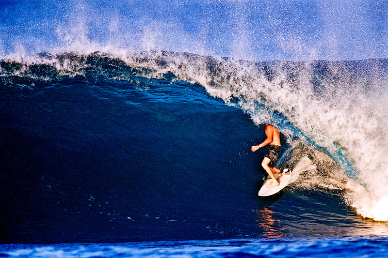 Surfer crouched inside a large barreling wave lit by golden evening light, spray fanning off the breaking crest