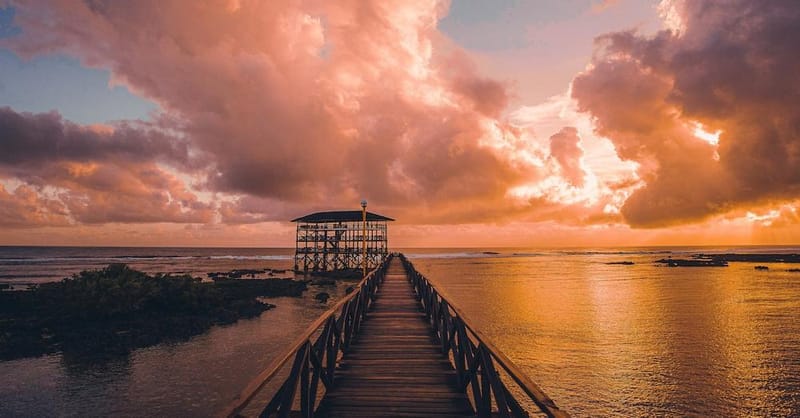 Wooden pier extending over calm turquoise lagoon water toward a thatched-roof viewing deck under a dramatic orange sunset sky in Siargao