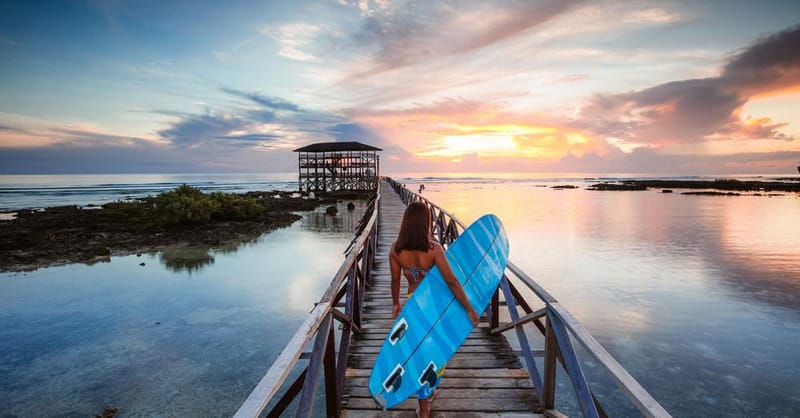 A surfer carrying a blue surfboard walks along a long wooden pier over calm water toward a thatched-roof deck at sunset in Siargao