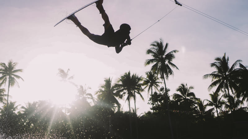 A wakeboarder flips upside down while catching big air against a bright sky, silhouetted with palm trees at Siargao Wakepark, General Luna