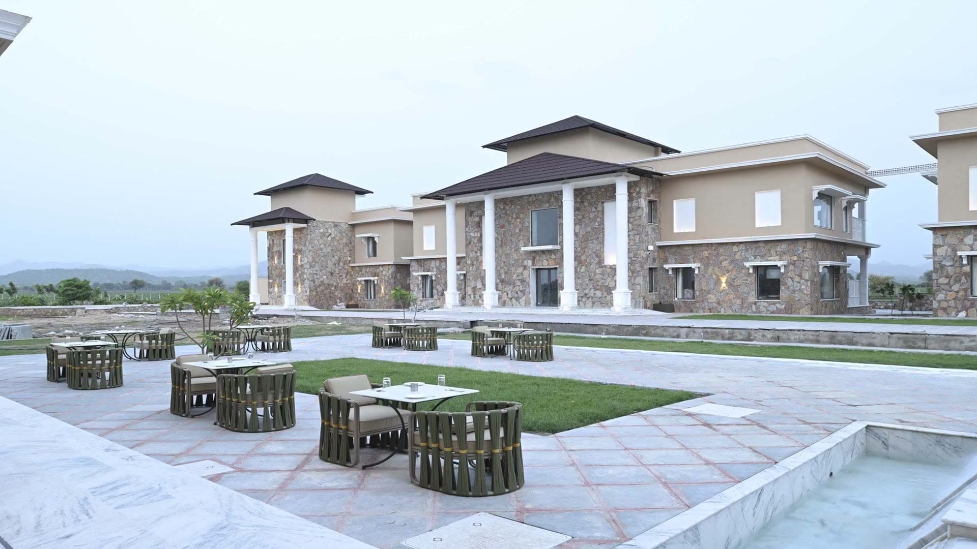 Outdoor dining tables on the terrace with a lawn, stone villas, and hills behind at dusk