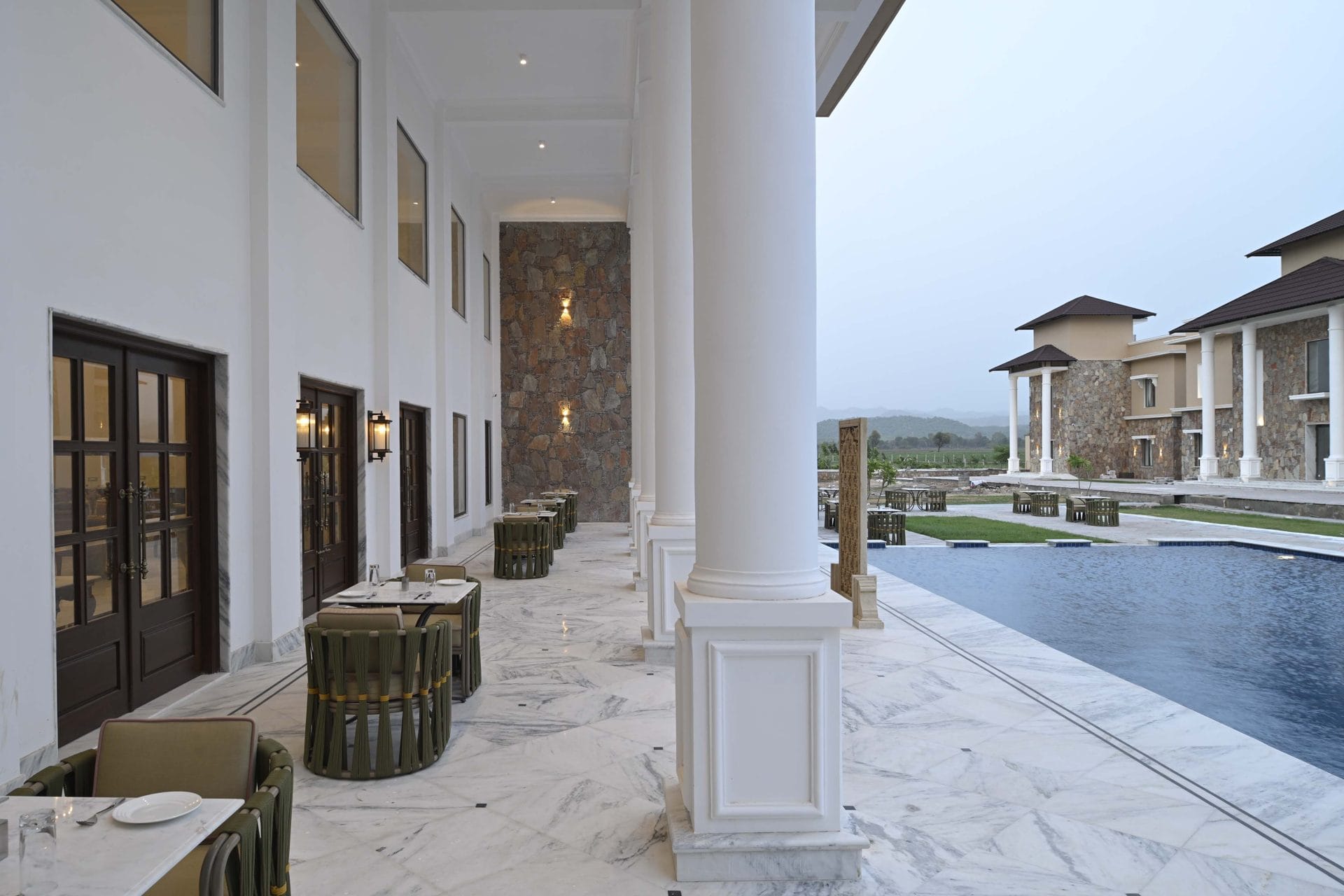 Dining tables on the marble colonnade beside the pool, villas, and hills behind at dusk